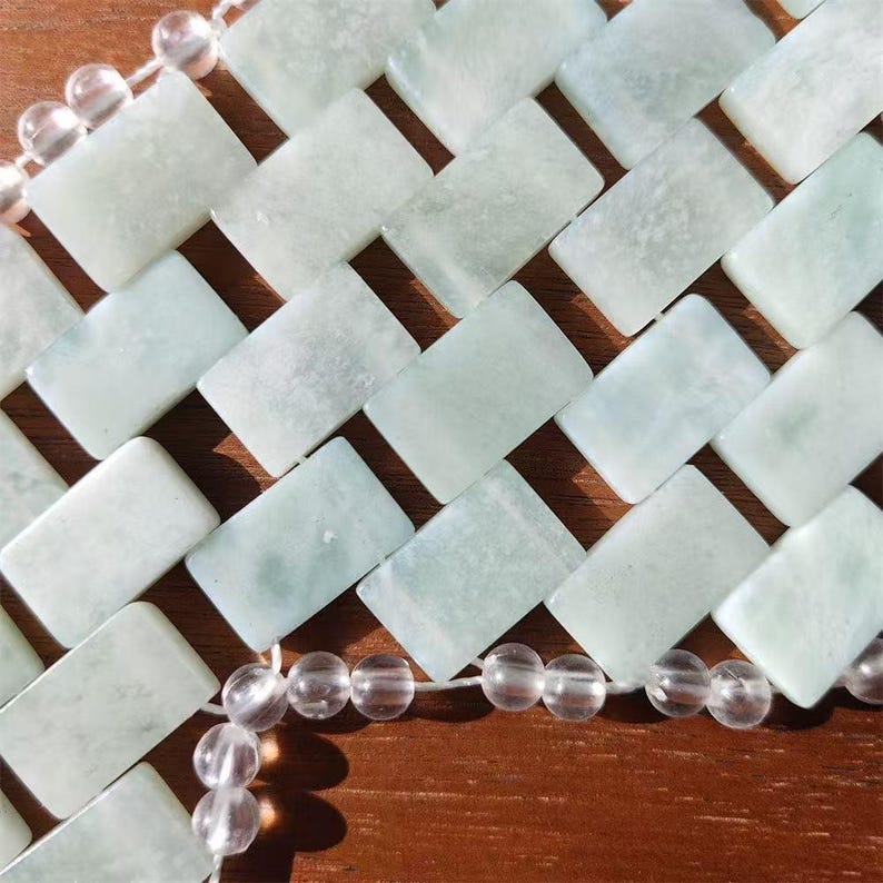 Puede incluir: Primer plano de una herramienta de masaje de jade. La herramienta est&aacute; compuesta por piedras rectangulares de jade verde p&aacute;lido dispuestas en un patr&oacute;n tejido. Cuentas redondas y transparentes est&aacute;n ensartadas a lo largo de los bordes. El fondo es una superficie de madera marr&oacute;n c&aacute;lida.