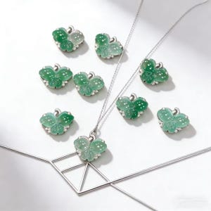 Puede incluir: Un conjunto de colgantes y pendientes de mariposa de jade verde con engastes de plata y peque&ntilde;as piedras blancas. Un colgante cuelga de una cadena de plata. Las joyas se exhiben sobre una superficie blanca.
