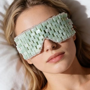 Puede incluir: Una m&aacute;scara de ojos de jade verde claro con un dise&ntilde;o tejido. La m&aacute;scara est&aacute; bordeada con peque&ntilde;as cuentas transparentes. La persona de la imagen est&aacute; acostada sobre una almohada blanca.