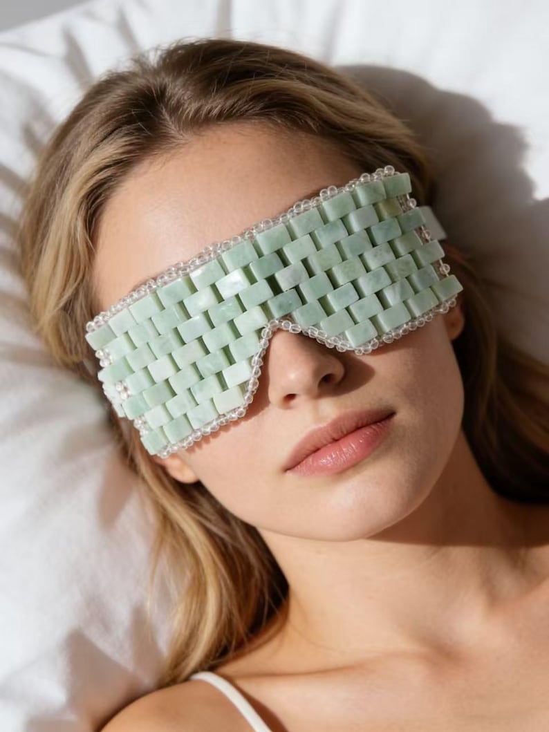 Puede incluir: Una m&aacute;scara de ojos de jade verde claro con un dise&ntilde;o tejido. La m&aacute;scara est&aacute; bordeada con peque&ntilde;as cuentas transparentes. La persona de la imagen est&aacute; acostada sobre una almohada blanca.