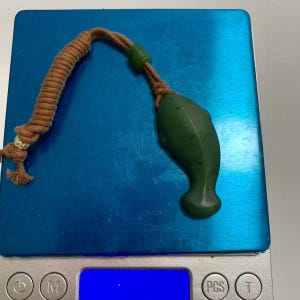 Puede incluir: Un colgante de delf&iacute;n de jade verde con un cord&oacute;n marr&oacute;n y una cuenta verde. El colgante est&aacute; sobre una balanza azul. El delf&iacute;n est&aacute; tallado con l&iacute;neas suaves y tiene una forma redondeada.