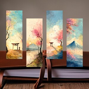 以下が含まれることがあります： 日本の風景を描いた4つの水彩画のブックマーク。各ブックマークは、鳥居、桜、山、夕日の異なるシーンを描いています。ブックマークは、茶色の革のジャーナルに表示されています。