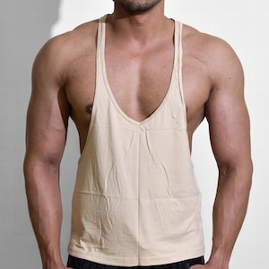 Puede incluir: Una camiseta sin mangas beige con cuello en V profundo. La camiseta sin mangas se combina con pantalones cortos deportivos negros. El modelo está de pie frente a un fondo blanco.