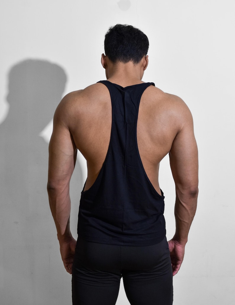 Puede incluir: Camiseta sin mangas negra con dise&ntilde;o de espalda deportiva. La parte superior sin mangas es usada por una persona. La persona lleva pantalones negros. El fondo es una pared blanca lisa. La camiseta tiene un dise&ntilde;o de espalda abierta.