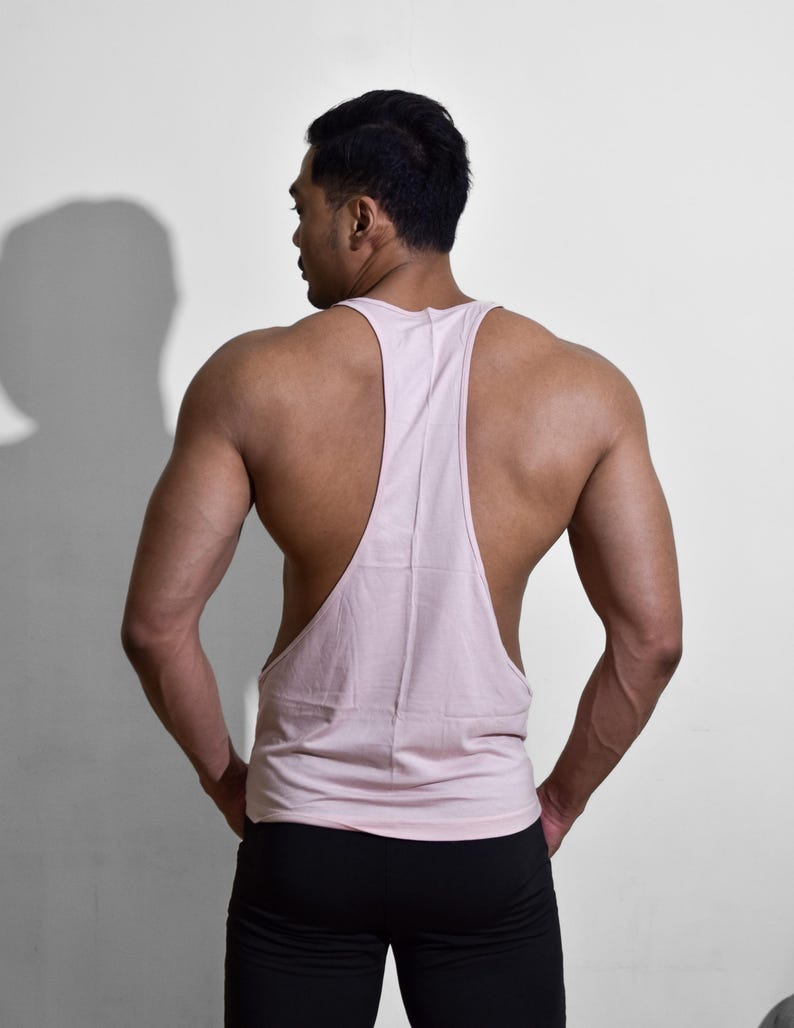 Puede incluir: Camiseta sin mangas rosa claro con dise&ntilde;o de espalda deportiva. La camiseta sin mangas se combina con pantalones deportivos negros. La persona est&aacute; de espaldas, con las manos en los bolsillos, sobre un fondo blanco.