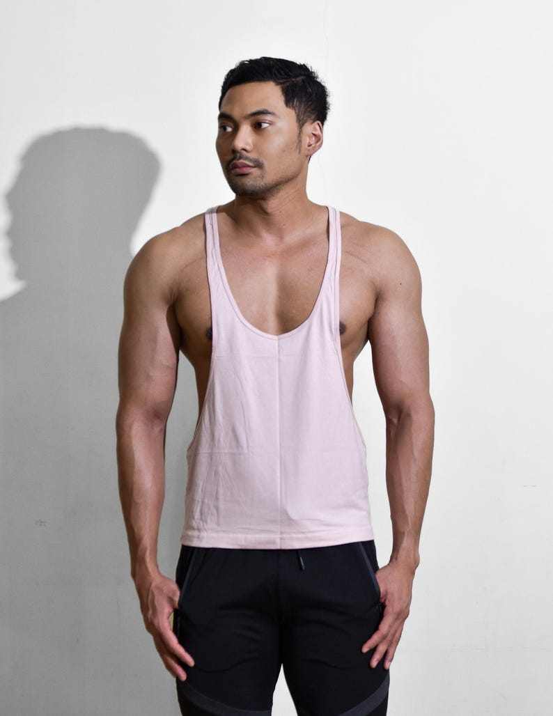 Puede incluir: Una camiseta sin mangas rosa claro con cuello redondo. La camiseta sin mangas es de material suave. El modelo lleva pantalones cortos deportivos negros. El fondo es una pared blanca lisa.
