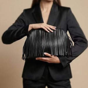Könnte beinhalten: Schwarze Clutch mit langen Fransen. Die Clutch wird von einer Person gehalten, die einen schwarzen Blazer und eine Hose trägt. Der Hintergrund ist einfarbig beige. Die Clutch ist der Mittelpunkt des Bildes.