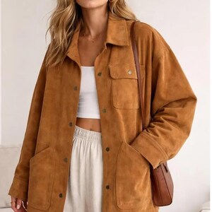 Könnte beinhalten: Eine hellbraune Jacke in Wildlederoptik mit durchgehender Knopfleiste, klassischem Kragen, langen Ärmeln, zwei großen aufgesetzten Vordertaschen und einer Brusttasche. Die Jacke wird über einem weißen bauchfreien Top und einer hellen Hose getragen.