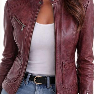 Könnte beinhalten: Burgunderfarbene Lederjacke mit Stehkragen, Reißverschlusstaschen und Frontreißverschluss. Die Jacke wird über einem weißen Oberteil und einer blauen Jeans getragen, mit einem schwarzen Gürtel mit goldener Schnalle.