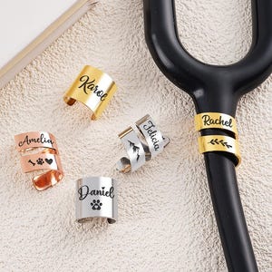 Puede incluir: Una colección de dijes de estetoscopio personalizados en oro, plata y oro rosa. Cada dije está grabado con un nombre, una huella de pata o un diseño de montaña. Los dijes se exhiben sobre una superficie blanca junto a un estetoscopio negro.