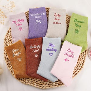Op de afbeelding: Een collectie van acht enkelsokken met glitterende texturen, elk voorzien van unieke geborduurde teksten en symbolen zoals My Happy Place, Transform & Glow, Ruby love, Dream Big, Shine Bright, Butterfly Soul, Mia darling en Stay Bright.