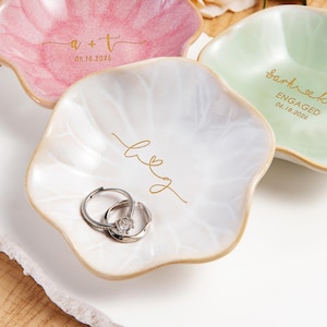 May include: Three flower-shaped ceramic trinket dishes in pink, white, and light green. The pink dish features gold text 'a + t' and '05.18.2026'. The white dish has gold text 'l & g'. The green dish has gold text 'sarh & kwin', 'ENGAGED', and '06.18.2026'. Two silver rings rest in the white dish.