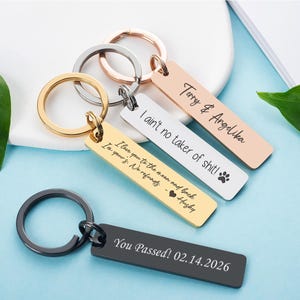 May include: A collection of personalized keychains in various colors, including gold, silver, rose gold, and black. Each rectangular keychain features engraved text, with messages like names, phrases, and a date. The keychains are attached to metal key rings.