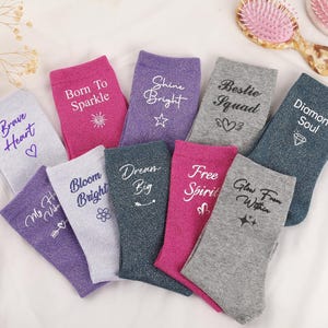 May include: A collection of ten assorted socks in shades of gray, purple, and pink, each featuring unique printed text and small graphic icons. The phrases include Brave Heart, Born To Sparkle, Shine Bright, Bestie Squad, Diamond Soul, and others.