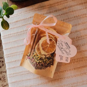 Op de afbeelding: Een doorzichtige cellofaan zak, vastgebonden met een roze lint, gevuld met gedroogde sinaasappelschijfjes, kaneelstokjes en kruiden. Een hartvormig label leest "Love Spell Simmer Pot". De zak ligt op een lichtroze gestreepte stof.