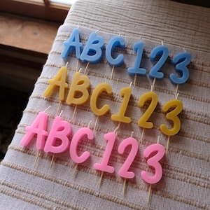 May include: Three rows of birthday candles in blue, yellow, and pink. Each row spells out "ABC123" in a bold, rounded font. The candles are on wooden sticks, set against a textured, beige fabric.