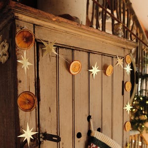 Puede incluir: Un armario de madera rústico adornado con una guirnalda de rodajas de naranja secas y adornos en forma de estrella. Un calcetín navideño verde y rojo cuelga de la puerta del armario, creando una escena festiva.