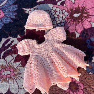May include: A light pink, handmade crochet baby dress and matching hat. The dress features a round neckline, short sleeves, and a flared skirt. The hat is a simple beanie style. The set is displayed on a floral patterned surface.