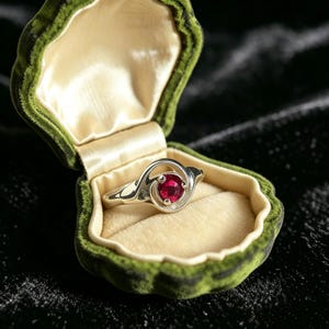 May include: A silver ring featuring a round, faceted red gemstone set in a swirling, open-work metal band. The ring is displayed inside an open, clam-shaped jewelry box with a dark green velvet exterior and a cream-colored satin interior lining.