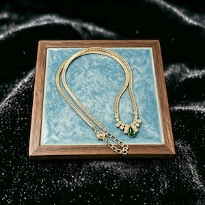 May include: A gold-toned necklace with a delicate chain and a pendant featuring a teardrop-shaped green gemstone. The necklace is displayed on a square, blue-tiled surface set within a wooden frame.