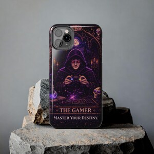 May include: A phone case featuring a digital illustration of a hooded figure playing a video game controller. The design includes a skull, candles, and the text "THE GAMER MASTER YOUR DESTINY." The case has a glossy finish and a dark purple color scheme.