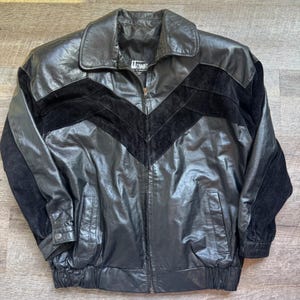 May include: Black leather jacket with a chevron design in black suede. The jacket has a collared neckline, a zipper closure, and two front pockets. The brand name "COMMIT" is visible on the label.
