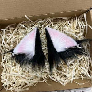 Può includere: Un paio di fermagli per capelli a forma di orecchie di gatto bianche e nere con interno rosa. Le orecchie in materiale soffice sono presentate in una scatola di cartone con imbottitura di carta.