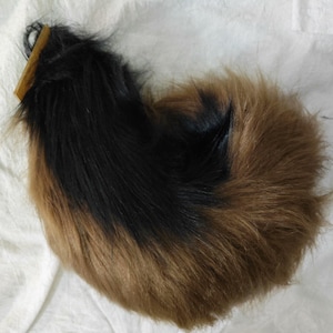 May include: A curled hairpiece with a gradient of colours. The top section is black, transitioning to a medium brown at the bottom. A wooden comb is attached at the top. The hairpiece is lying on a white surface.