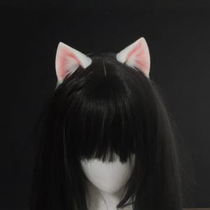 Puede incluir: Una peluca negra con pelo largo y liso y flequillo, adornada con orejas de gato blancas con interior rosa. Las orejas están colocadas en la parte superior de la cabeza. El fondo es negro, resaltando el contraste de las orejas blancas y rosas.