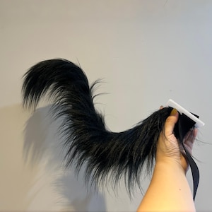 May include: A fluffy black tail with a white and black attachment. The tail is held by a black strap and is made of long, soft, and textured material. The tail is curved and has a natural appearance.