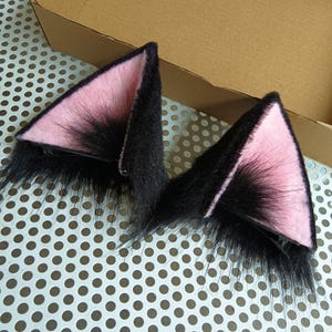 May include: A pair of triangular cat ear hair clips featuring long black faux fur on the exterior and a soft light pink fabric lining on the interior. Each ear is attached to a black metal alligator hair clip for secure attachment to hair.