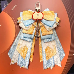 May include: A decorative ribbon rosette featuring layered yellow and light blue patterned fabric strips, topped with a red apple-shaped charm and a silver-toned metal clasp. A small silver airplane charm hangs from a chain in the center.
