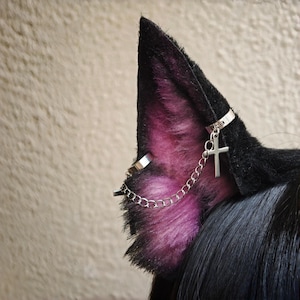 Può includere: Un paio di orecchie da animale in pelliccia sintetica con pelo esterno nero e pelo interno viola vivace. Le orecchie sono decorate con anelli in metallo argentato collegati da una catenina, con un piccolo ciondolo a croce argentato.