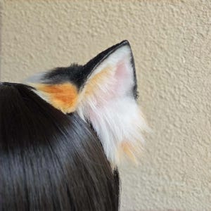 Puede incluir: Un par de orejas de gato de piel sintética en negro, blanco y naranja. Las orejas están unidas a una diadema negra y están colocadas sobre cabello oscuro. El interior de la oreja es de color rosa claro.