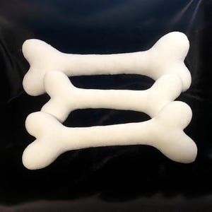 May include: Three plush, white, bone-shaped toys arranged in a stacked formation. Each toy features a smooth, rounded surface with the classic two-lobed ends characteristic of a bone shape, presented against a dark, solid background.