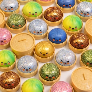 May include: A collection of colorful, smiling dumpling-shaped bath bombs in small, round, bamboo-style containers. The bath bombs are various colors, including blue, green, gold, pink, and silver, with glittery and solid finishes. The containers have lids.