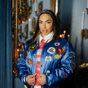 Puede incluir: Chaqueta bomber azul satinada con varios parches bordados, incluyendo corazones y texto. La chaqueta se lleva sobre una camisa roja con el texto "GID * U". La persona lleva una camisa blanca con cuello y vaqueros azules.