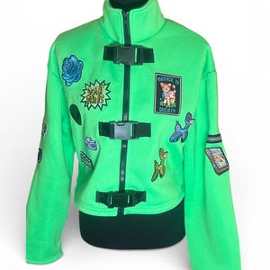 Può includere: Giacca verde brillante con cerniera nera, fibbie e toppe. Le toppe includono una rosa blu, le parole "DON'T PANIC", un cervo con la scritta "MENACE TO SOCIETY", una fotocamera e un animale a palloncino. La giacca ha un collo alto.