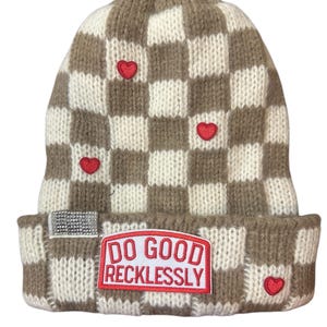 Op de afbeelding: Een bruin-witte geruite beanie met rode hartaccenten. De beanie heeft een rechthoekige patch met de tekst "DO GOOD RECKLESSLY" en een kleine rechthoekige strass-patch.