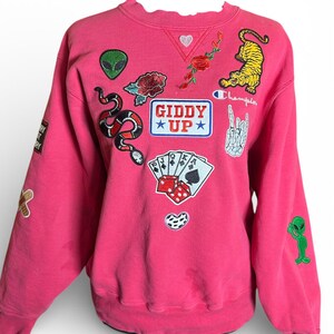 Puede incluir: Una sudadera rosa Champion adornada con varios parches bordados. Los parches incluyen un tigre, un extraterrestre, una serpiente, rosas, naipes, dados y el texto "GIDDY UP". La sudadera tiene cuello redondo y mangas largas.