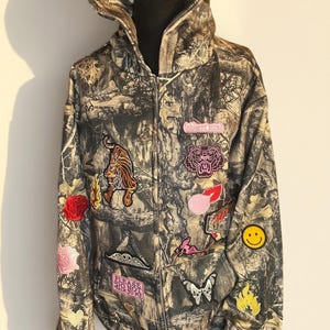 Könnte beinhalten: Ein Camouflage-Kapuzenpullover mit Reißverschluss und verschiedenen gestickten Aufnähern. Zu den Aufnähern gehören ein Tiger, eine Rose, ein Smiley und der Text "PLEASE DO NOT TALK TO ME" und "(oh)sent intnt". Der Hoodie wird auf einer schwarzen Schaufensterpuppe präsentiert.
