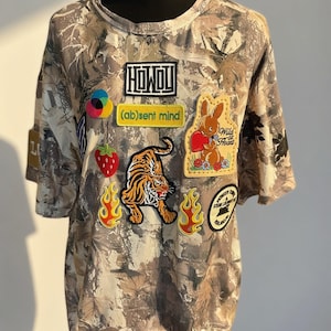 Puede incluir: Una camiseta de manga corta con un patrón de camuflaje de hojas en marrón y gris, decorada con varios parches bordados que incluyen un tigre, un conejo, llamas, una fresa y el texto HOWDY, (ab)sent mind y Wild at Heart.