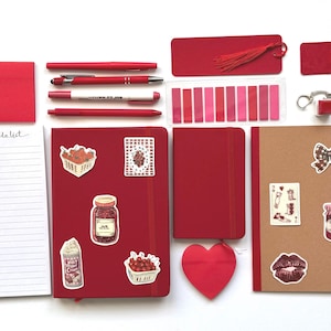 Puede incluir: Una colección de artículos de papelería rojos, que incluyen cuadernos, bolígrafos, notas adhesivas y un marcapáginas con borla. Pegatinas decorativas con diseños de comida y lazos están en los cuadernos. También se incluye un recorte en forma de corazón.