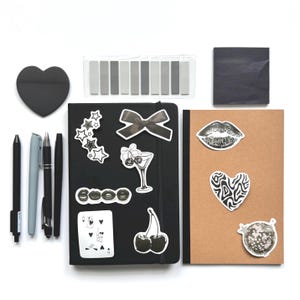 Puede incluir: Una colección de artículos de papelería, que incluye bolígrafos negros y grises, una nota adhesiva en forma de corazón, un cuaderno negro con pegatinas, un cuaderno marrón y pestañas adhesivas grises. Las pegatinas presentan varios diseños, como estrellas, labios y cerezas.