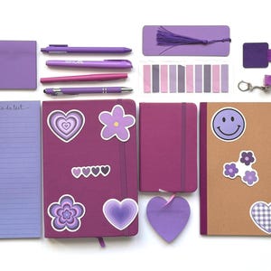 Puede incluir: Colección de artículos de papelería morados, que incluyen cuadernos, bolígrafos, notas adhesivas y un marcapáginas con borla. Los cuadernos presentan pegatinas de corazón y flores, y uno tiene una carita sonriente. Sobre fondo blanco.