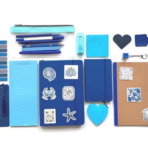 Puede incluir: Un surtido de artículos de papelería en tonos azules y turquesas, que incluyen cuadernos, bolígrafos, notas adhesivas y un estuche. Pegatinas decorativas con diseños de conchas marinas y corales adornan los cuadernos. También se ven un marcapáginas y un bloc de notas con el texto "to do list".