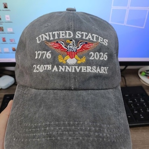Puede incluir: Gorra de b&eacute;isbol gris con texto bordado y un dise&ntilde;o de &aacute;guila patri&oacute;tica. El texto dice "UNITED STATES 1776 2026 250TH ANNIVERSARY". La gorra tiene una visera curva y un cierre de correa de tela.