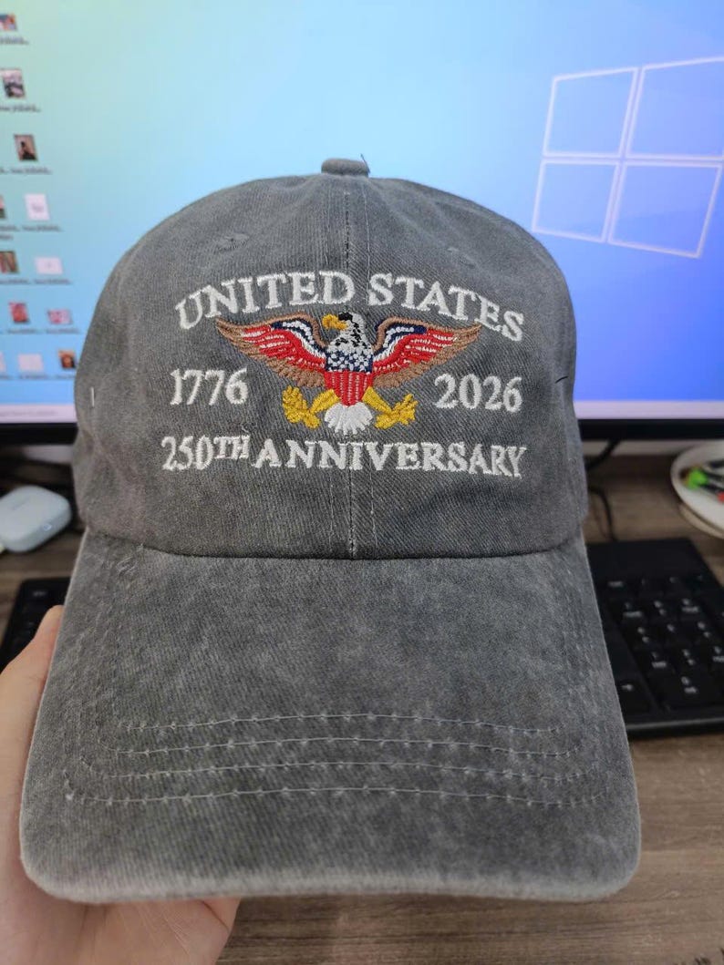 Puede incluir: Gorra de b&eacute;isbol gris con texto bordado y un dise&ntilde;o de &aacute;guila patri&oacute;tica. El texto dice "UNITED STATES 1776 2026 250TH ANNIVERSARY". La gorra tiene una visera curva y un cierre de correa de tela.