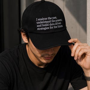 Op de afbeelding: Zwarte baseball cap met witte geborduurde tekst: "Ik analyseer het verleden, begrijp het heden en bouw datagestuurde strategieën voor de toekomst." Gedragen door een persoon in een zwart t-shirt.