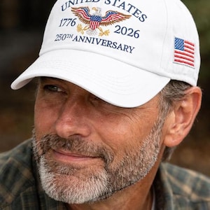 Puede incluir: Gorra de b&eacute;isbol blanca con el texto "UNITED STATES 1776 2026 250TH ANNIVERSARY" y un &aacute;guila. La gorra tambi&eacute;n presenta una peque&ntilde;a bandera estadounidense en el lateral. Un accesorio patri&oacute;tico cl&aacute;sico.